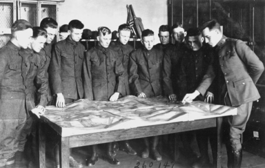 Military cadets using a topographic map in class, 1919. The image is a black and white photograph of eleven young men standing around a table with a model topographic map set upon it, all of them wearing cadet uniforms.